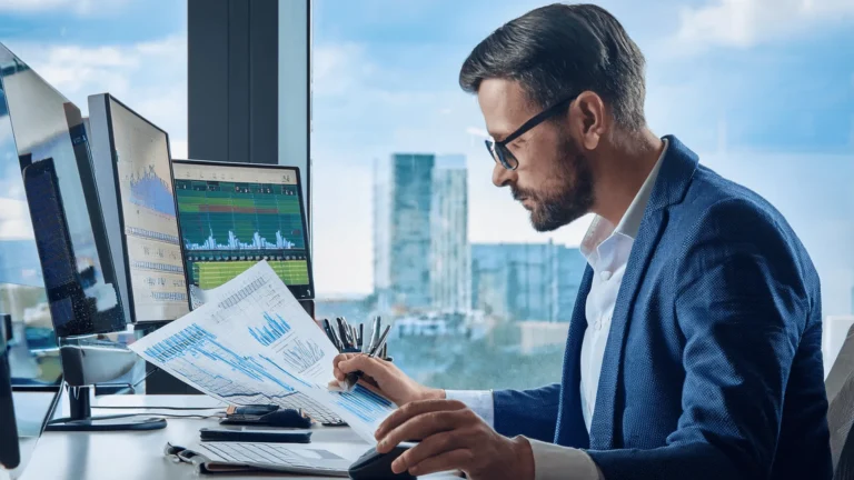 Mężczyzna w niebieskiej marynarce i okularach analizujący wykresy finansowe na ekranach komputerów i dokumentach w nowoczesnym biurze.