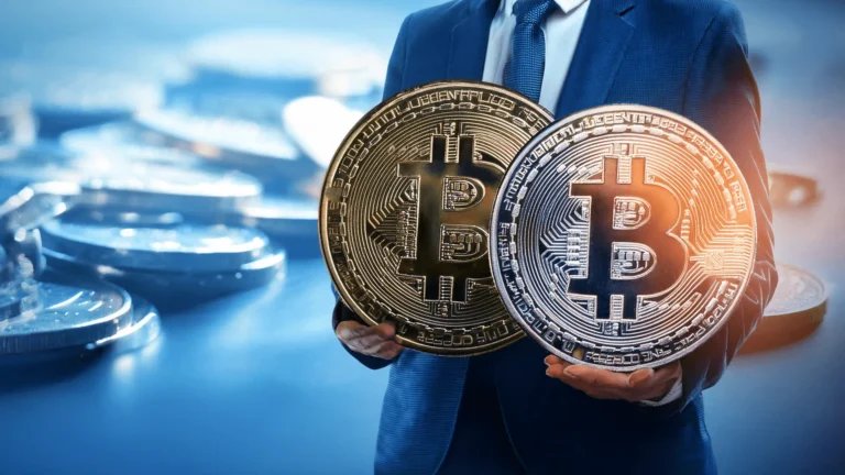 Mężczyzna w niebieskim garniturze trzymający dwie duże fizyczne monety Bitcoin: złotą i srebrną.