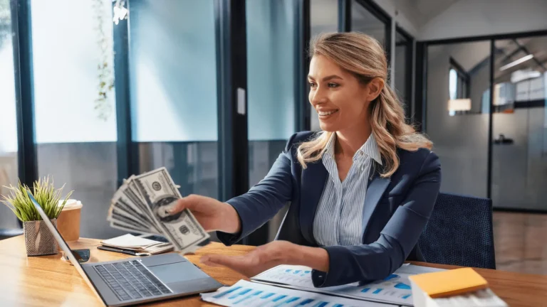 Uśmiechnięta kobieta w biurze trzymająca w dłoni wachlarz banknotów dolarowych, siedząc przy biurku z laptopem.