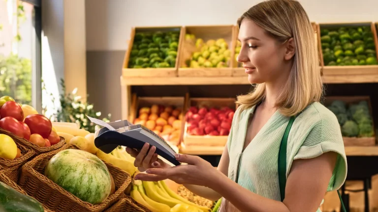 Młoda kobieta płacąca kartą za pomocą terminala płatniczego w sklepie ze świeżymi owocami i warzywami.