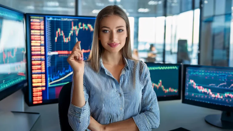 Młoda kobieta w paski koszuli uśmiecha się na tle monitorów z wykresami giełdowymi w nowoczesnym biurze finansowym.