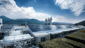 Nowoczesna fabryka z zainstalowanymi panelami słonecznymi na dachu, otoczona zielenią, z widokiem na góry w oddali.