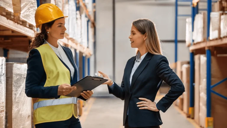 Dwie profesjonalne kobiety rozmawiające w magazynie, jedna w kasku ochronnym i kamizelce odblaskowej z podkładką do pisania.